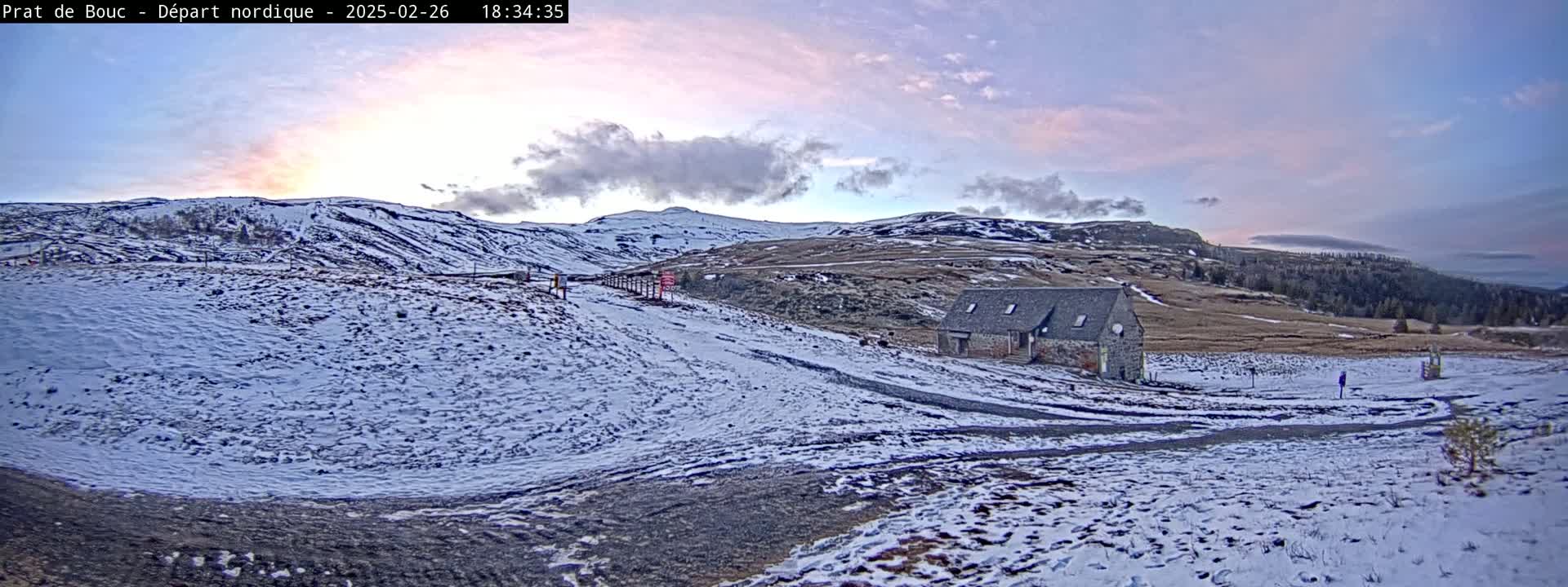 A snow-covered landscape at sunset, featuring a small stone building near a path leading up a hillside under a partly cloudy sky.
