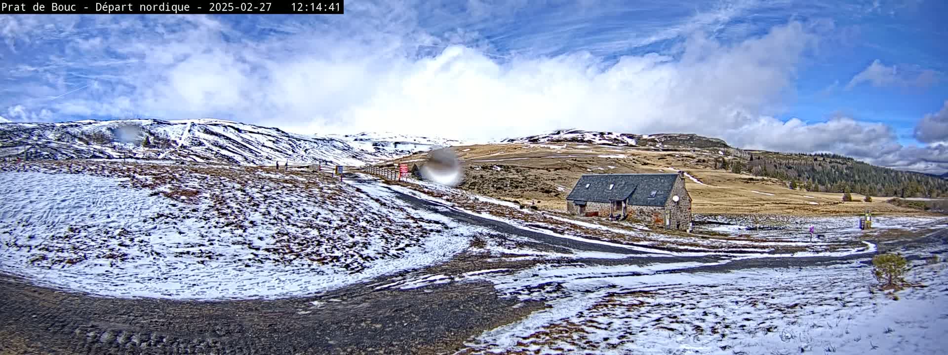 A stone building sits on a snow-dusted road amidst rolling hills partially covered in snow under a partly cloudy sky.