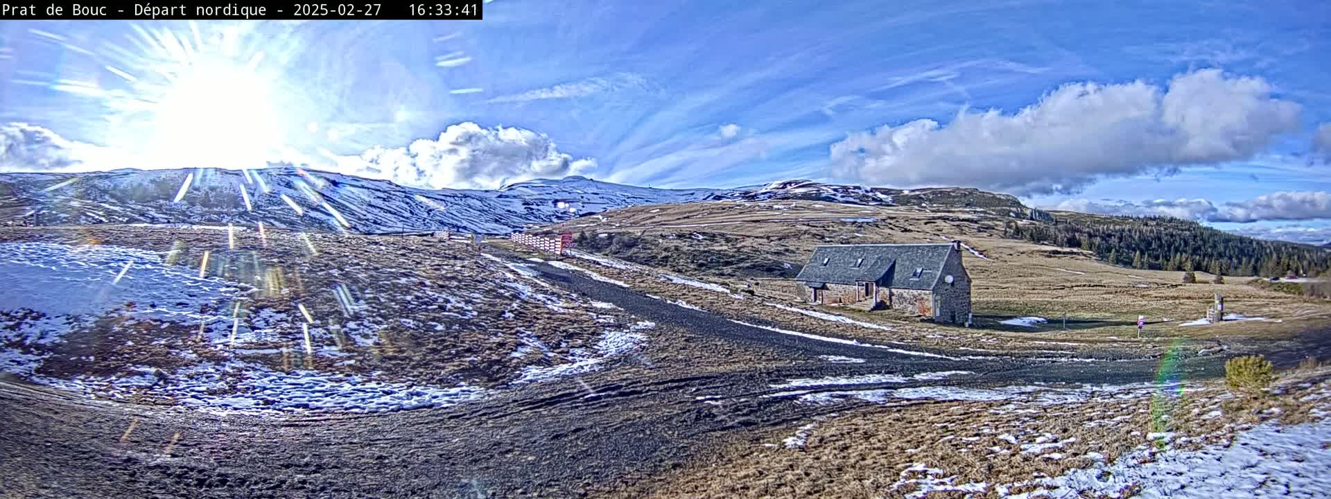 A sunny day with partly cloudy skies shows a snow-dusted landscape with a stone building and a road winding through the hills.