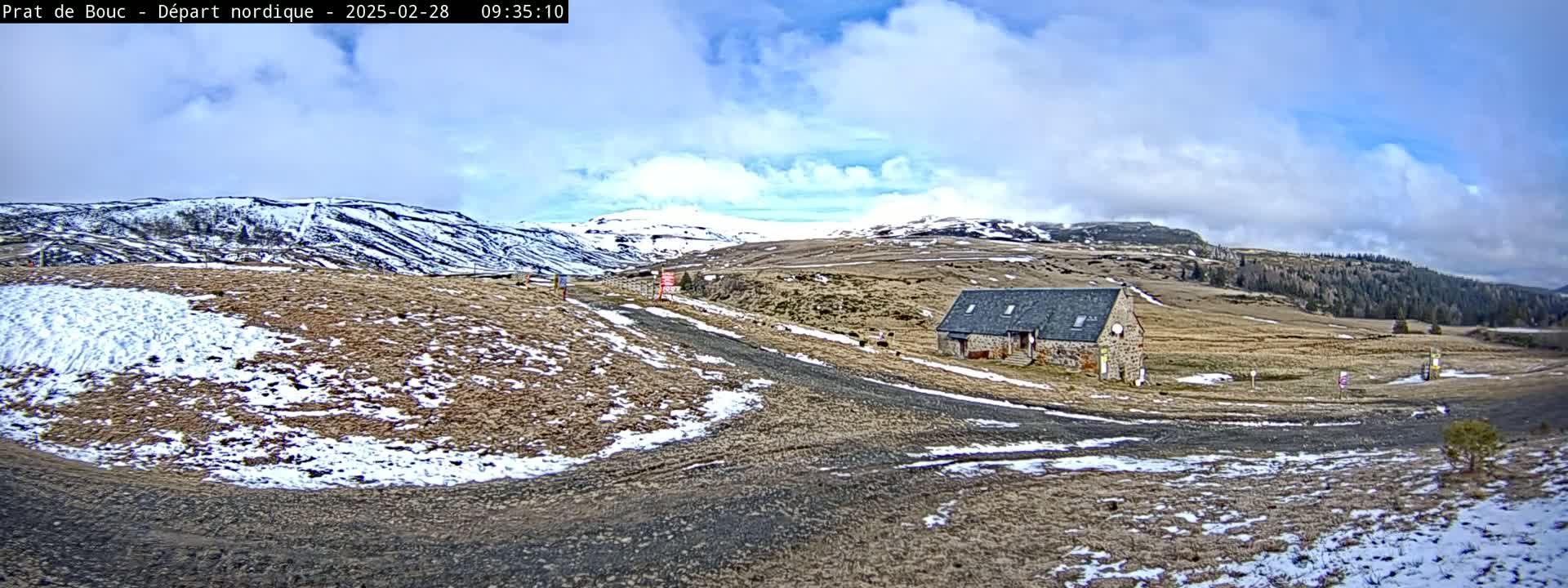 A stone building sits on a snow-dusted, grassy plateau, with snow-capped mountains in the background under a partly cloudy sky.