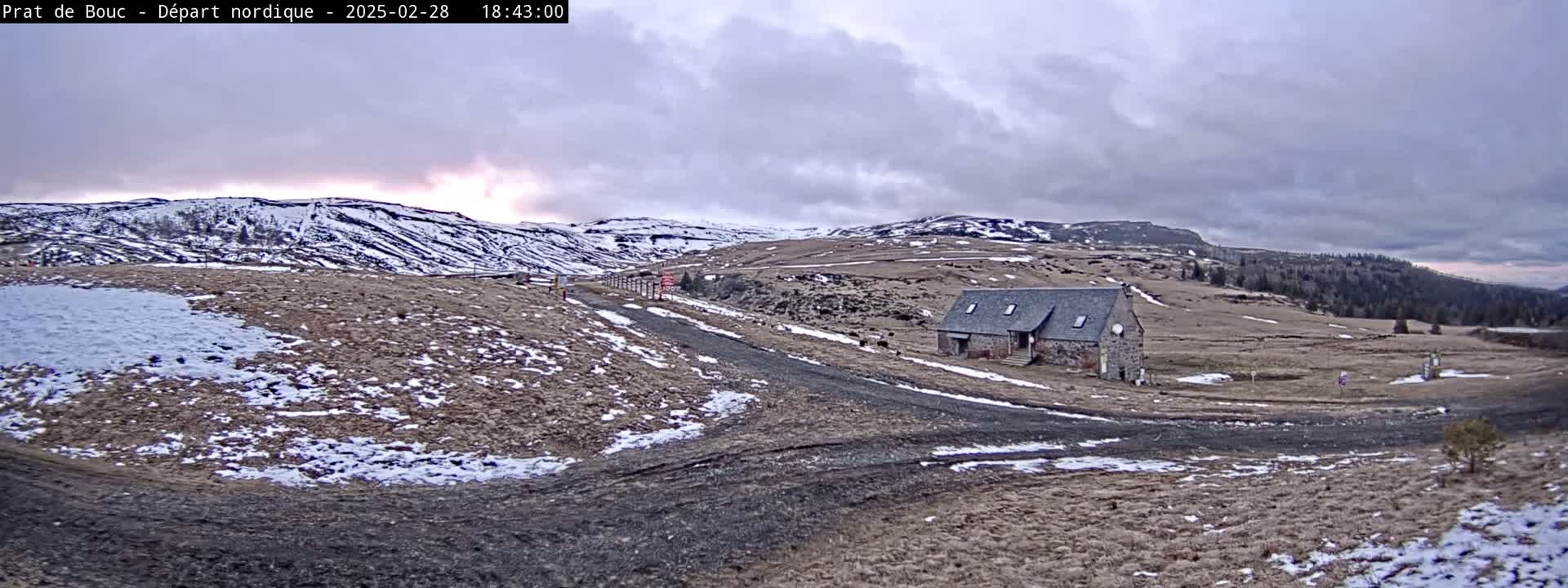 A long, low stone building sits on a snow-dusted, grassy plain beneath a mostly cloudy sky.