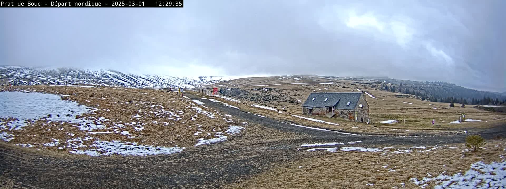 A stone building sits on a snow-dusted, grassy plateau under a cloudy sky.
