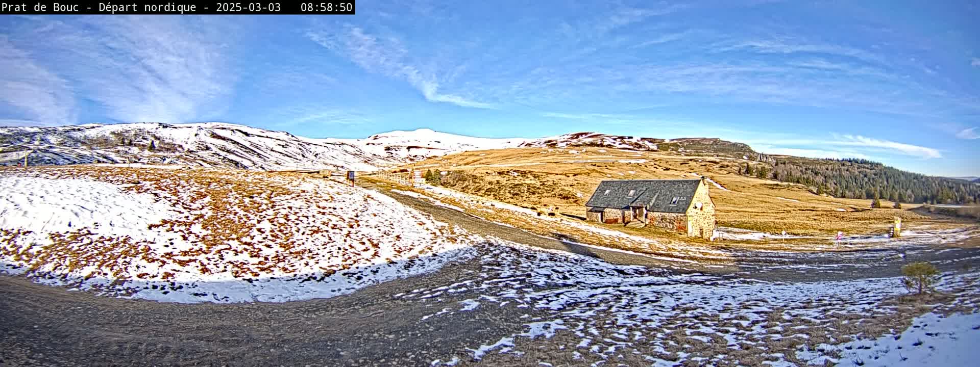 A stone building sits on a snow-dusted, brown hillside under a mostly clear blue sky.