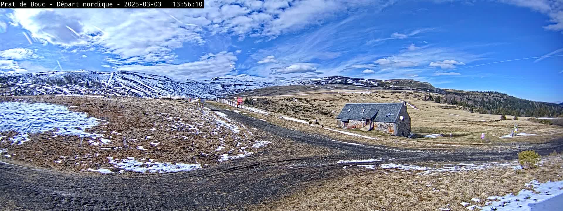 A stone building sits on a dirt road in a mountainous region with patches of snow on the ground under a partly cloudy blue sky.