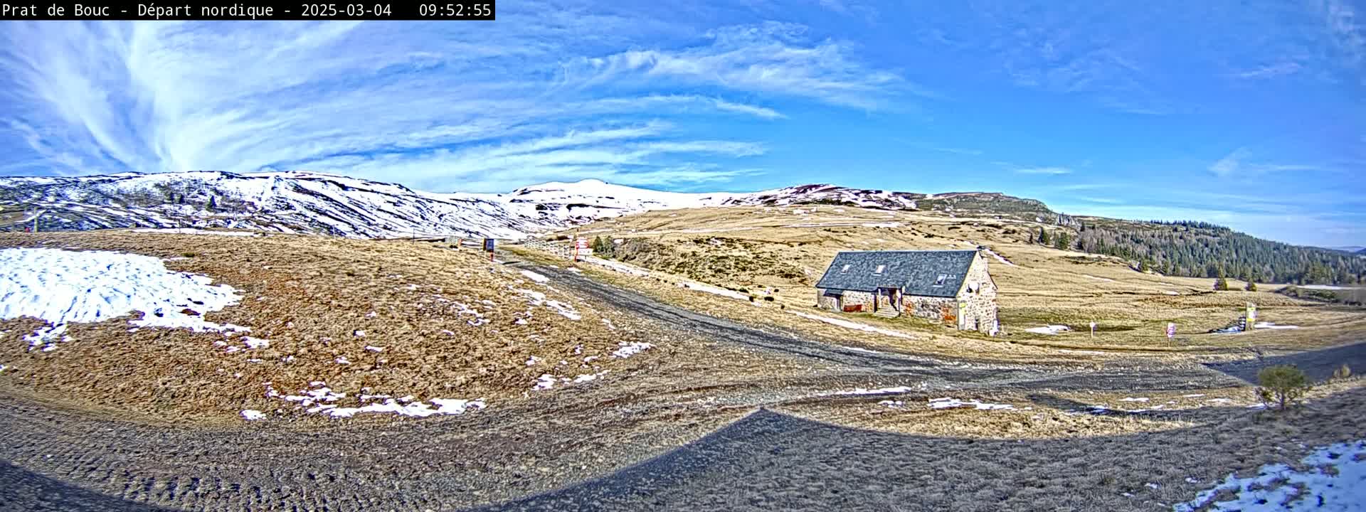 A stone building sits on a dirt road in a high-altitude landscape with patches of snow under a mostly clear blue sky.
