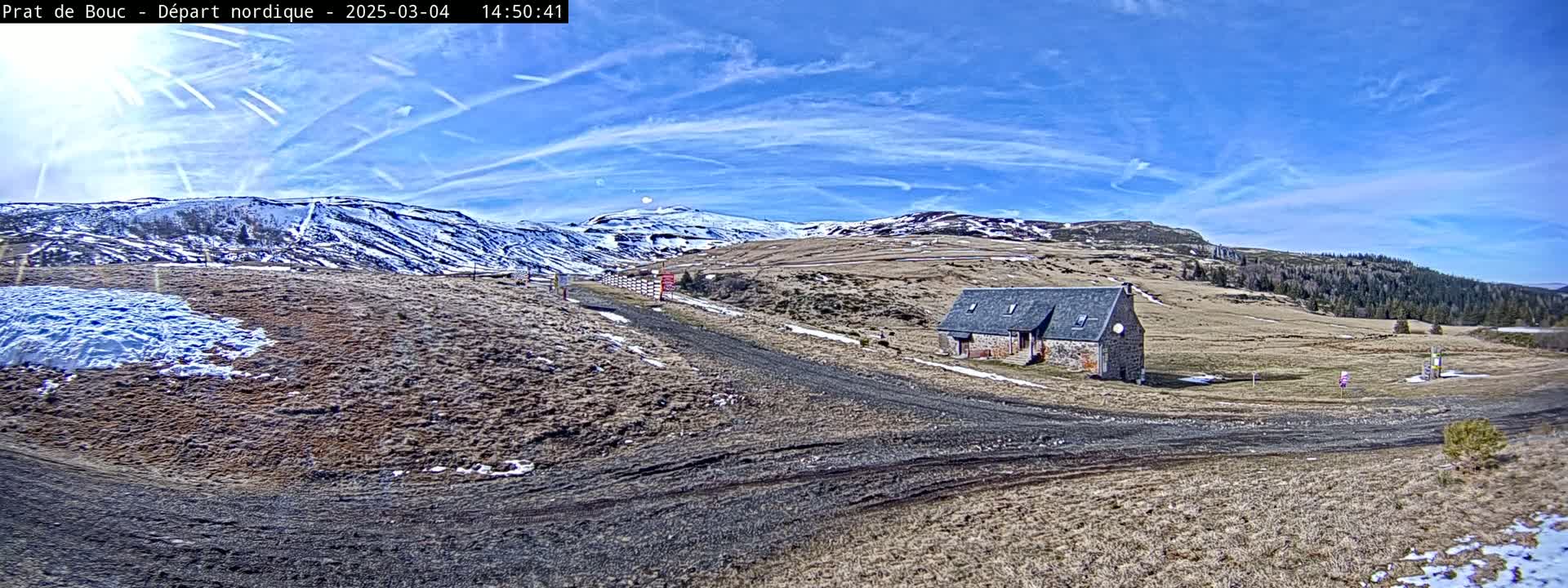 A stone building sits on a dirt road in a snow-dusted mountainous landscape under a mostly sunny sky with contrails.