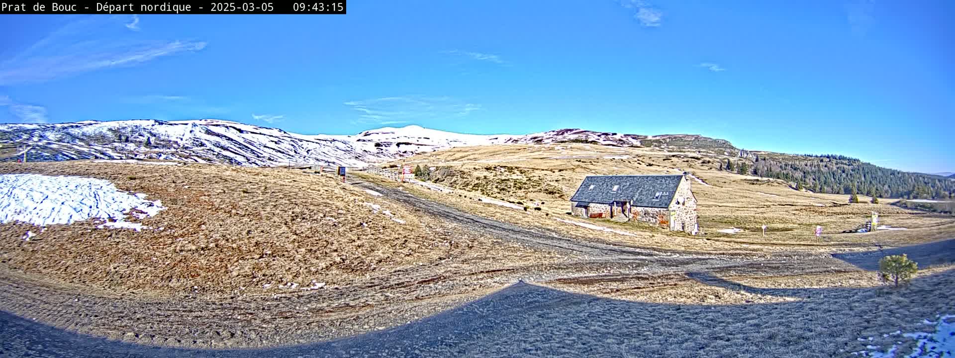 A stone building sits on a brown, snow-dusted hillside under a clear blue sky.