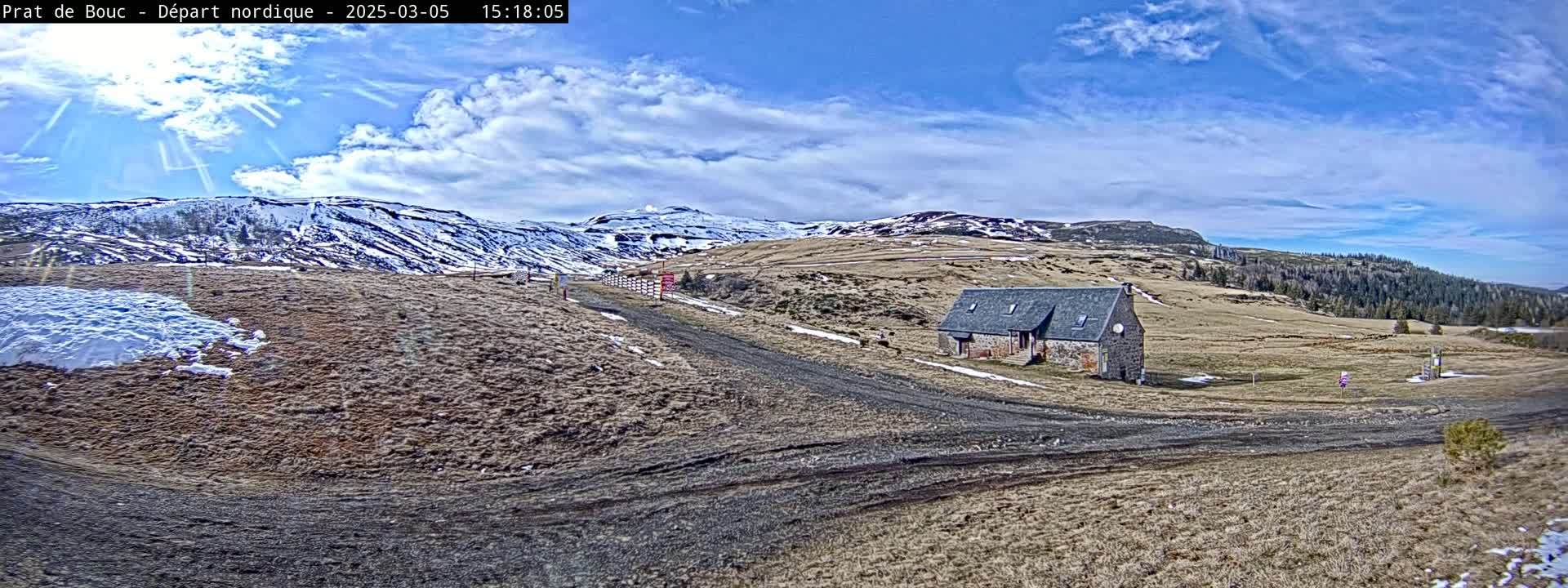 A stone building sits on a brown, grassy plateau with a dirt road leading to it, partially snow-covered hills in the background under a mostly sunny sky with some clouds.