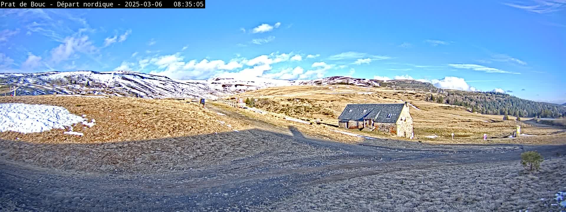 A stone building sits on a brown, snow-dusted hillside under a mostly sunny sky, with snow-capped mountains in the background.