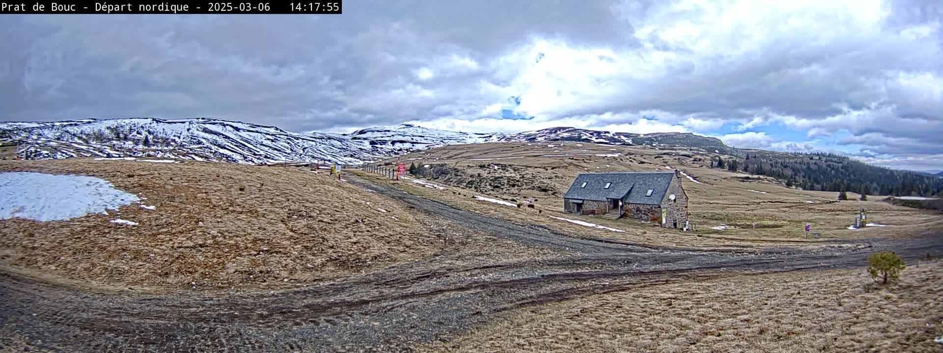 A stone building sits on a dirt road in a mountainous landscape with patchy snow and a cloudy sky.