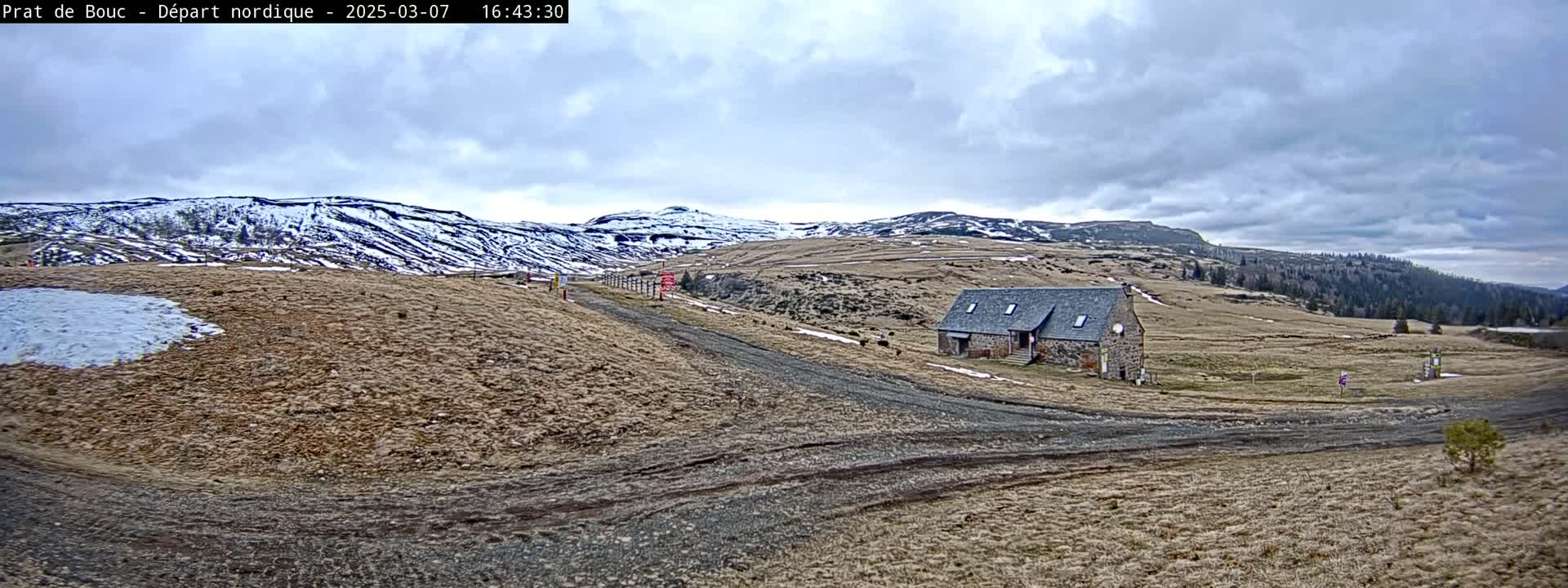 A stone building sits on a dirt road in a sparsely vegetated landscape under a cloudy sky, with patches of snow visible on nearby hills.