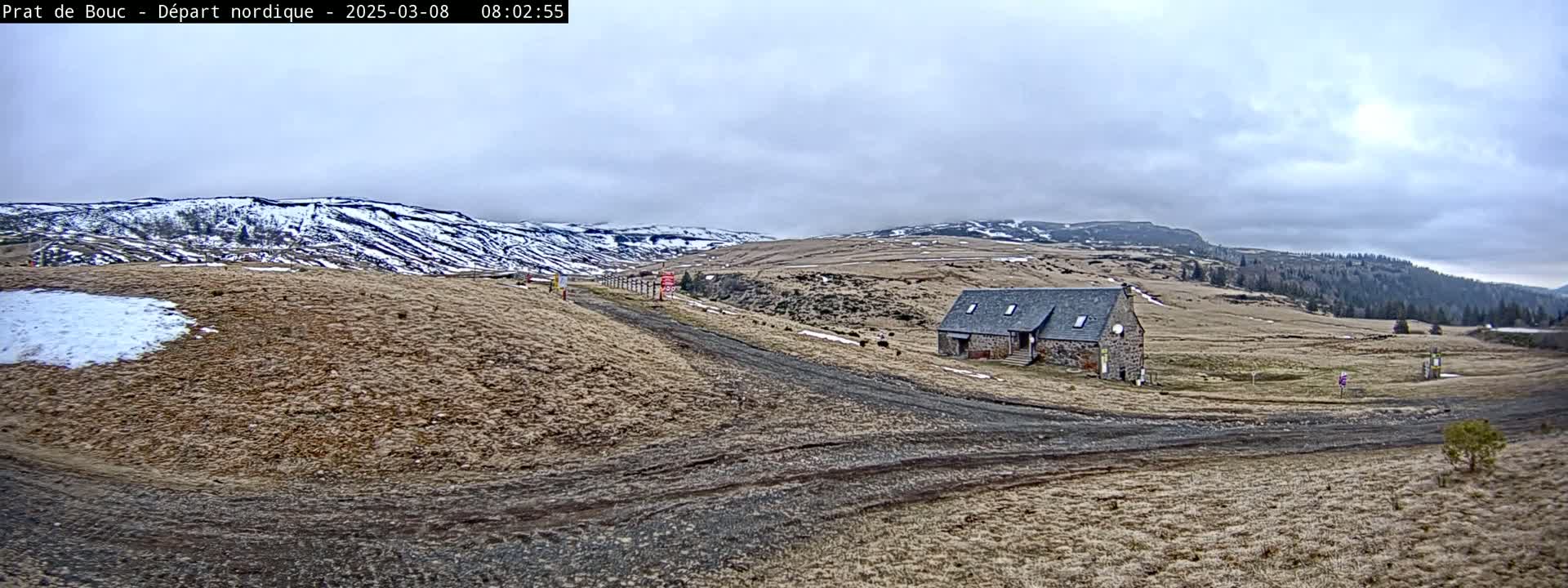 A stone building sits on a brown, grassy plain with a dirt road leading to it, under an overcast sky, with snow-capped hills in the background.