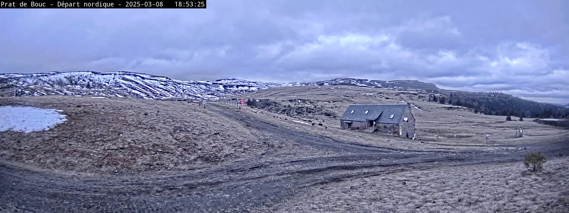 A stone building sits on a dirt road in a sparsely vegetated landscape under a cloudy sky, with snow dusting the distant hills.