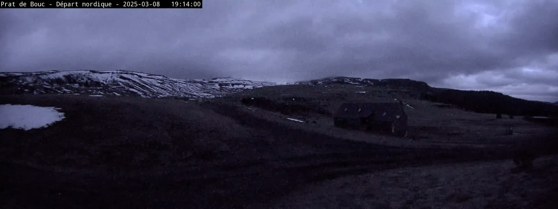 A dark, overcast sky hangs over a snow-dusted landscape featuring a small building near a hill, with patches of snow visible on the ground.