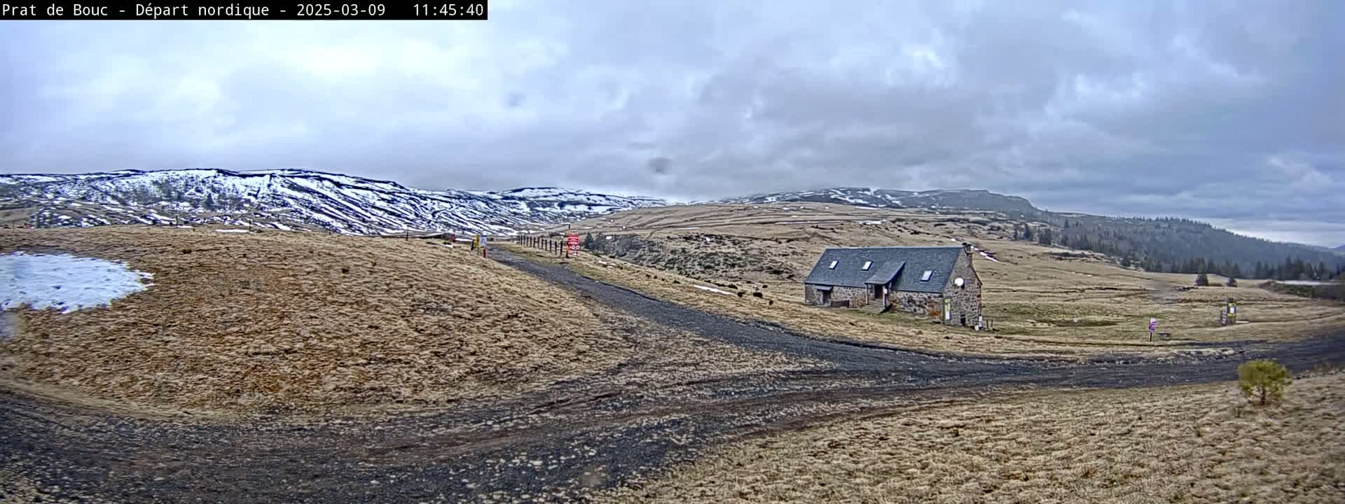 A stone building sits on a dirt road in a hilly, snow-dusted landscape under a cloudy sky.