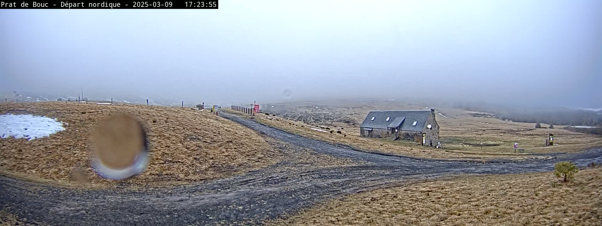 A stone building sits on a foggy, dry, brown hillside with a dirt road leading to it, and a small patch of snow is visible in the foreground.