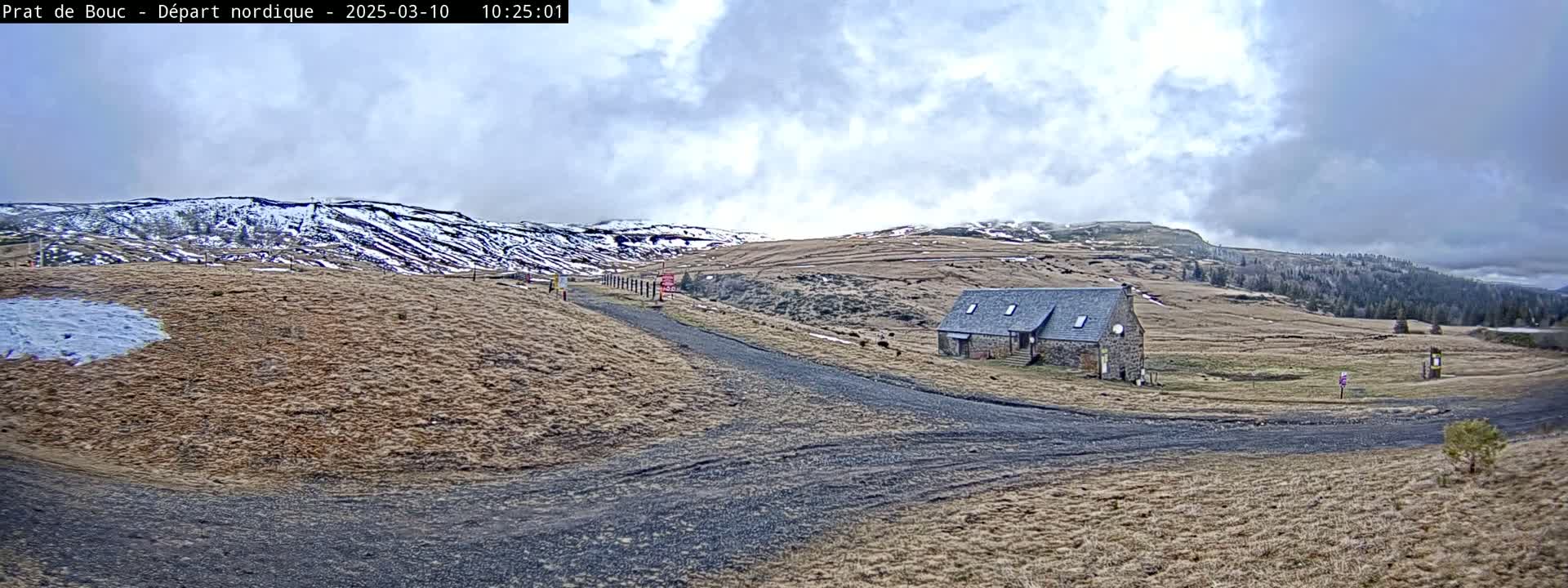 A stone building sits on a dirt road in a dry, grassy field under a cloudy sky, with snow-capped hills in the background.