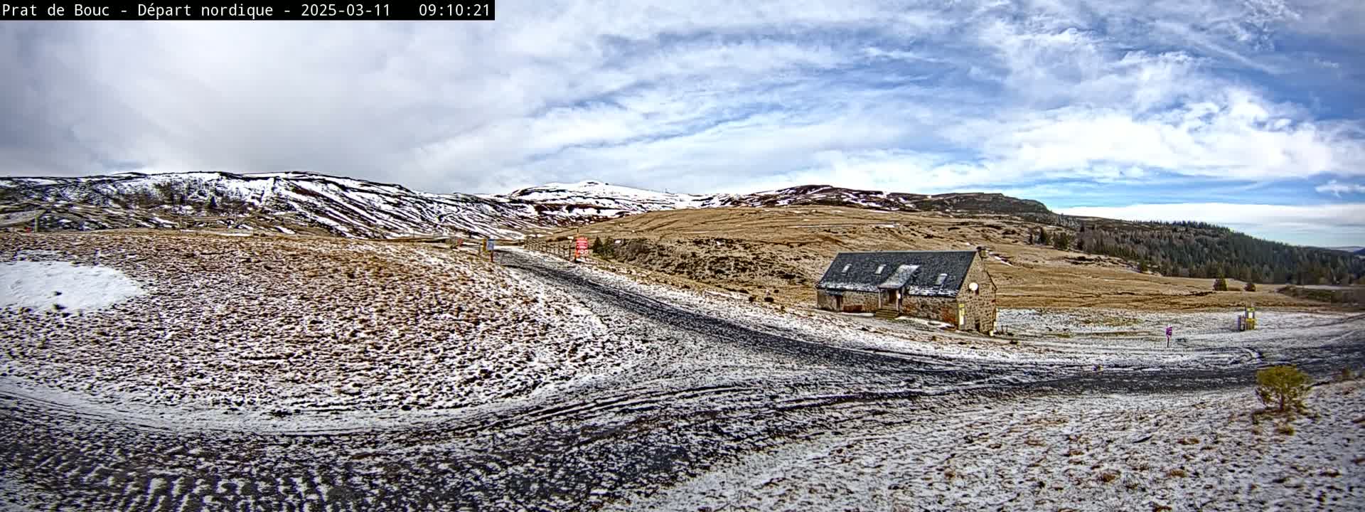A dirt road winds through a snow-dusted landscape towards a stone building nestled among brown hills under a partly cloudy sky.