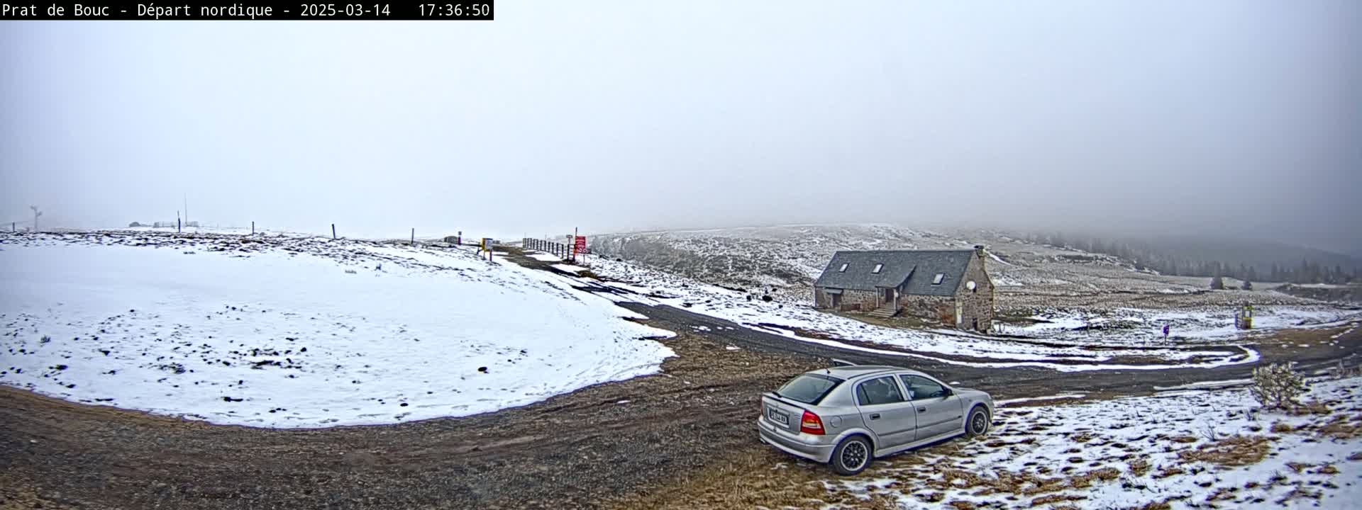 A silver car is parked near a stone building on a snow-covered road in foggy conditions.