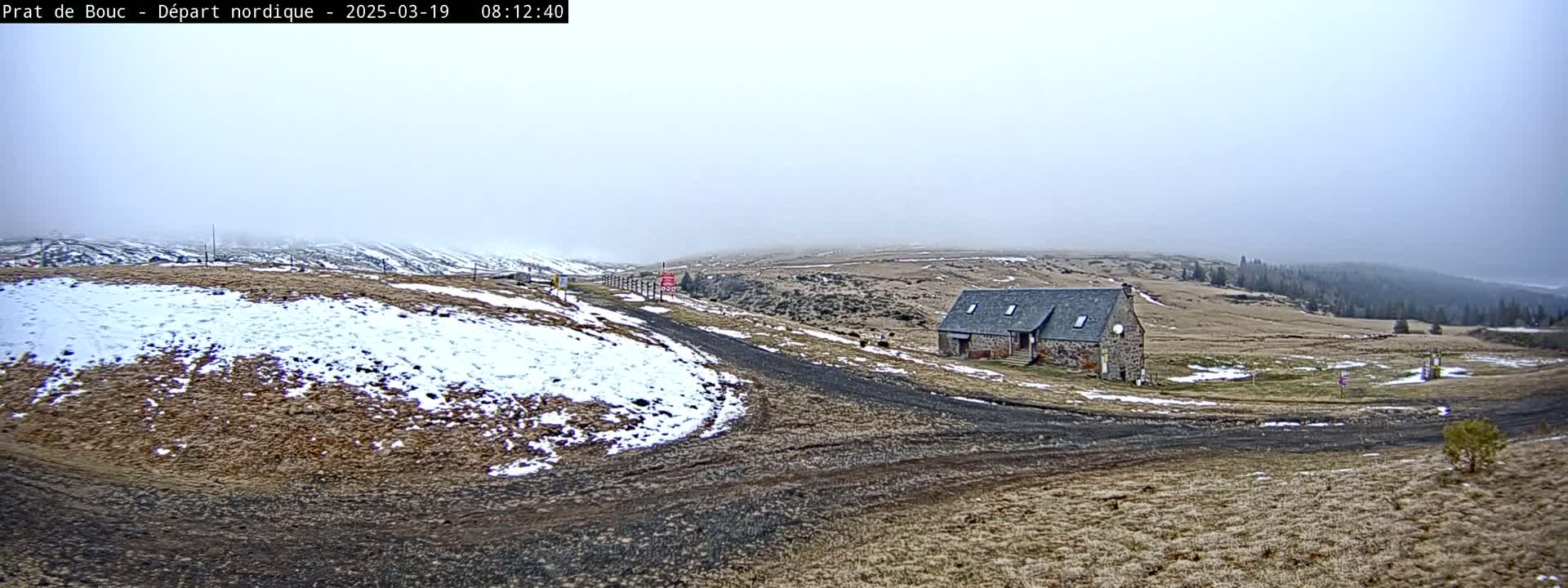 A stone building sits on a sparsely vegetated hillside with patches of snow under a foggy sky.