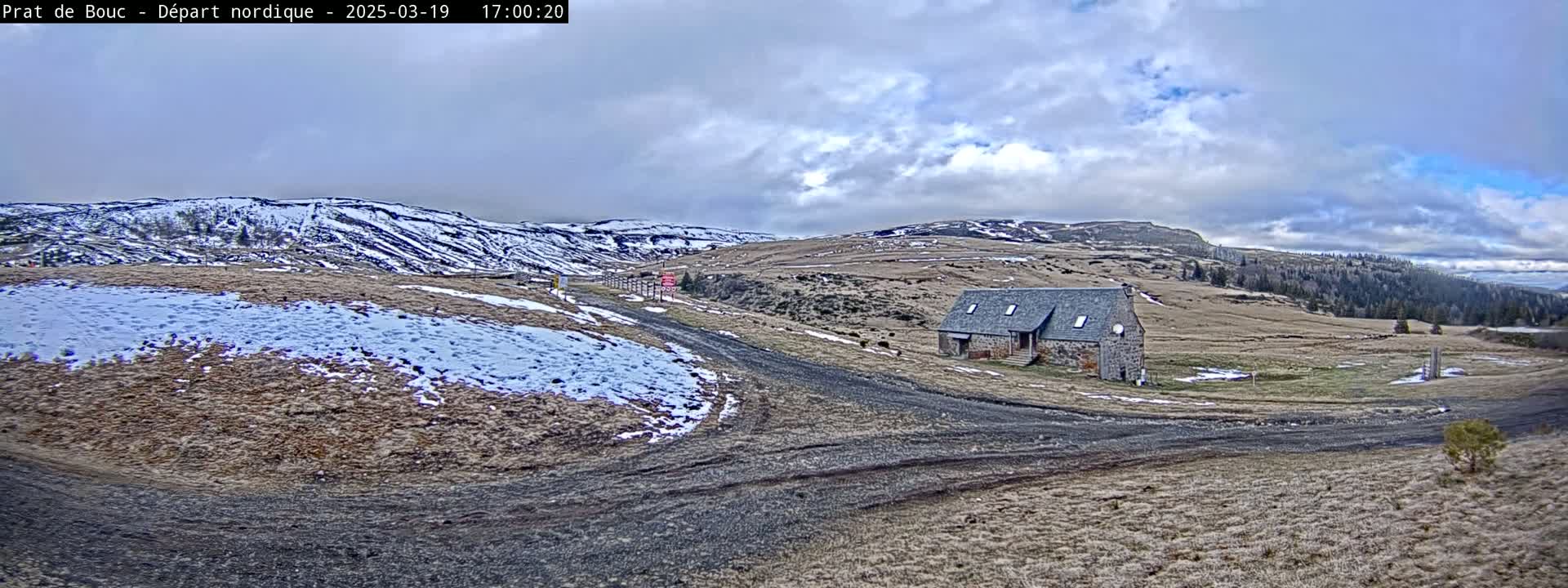A stone building sits on a dirt road in a sparsely vegetated landscape under a partly cloudy sky, with patches of snow on the ground and distant snow-capped hills.