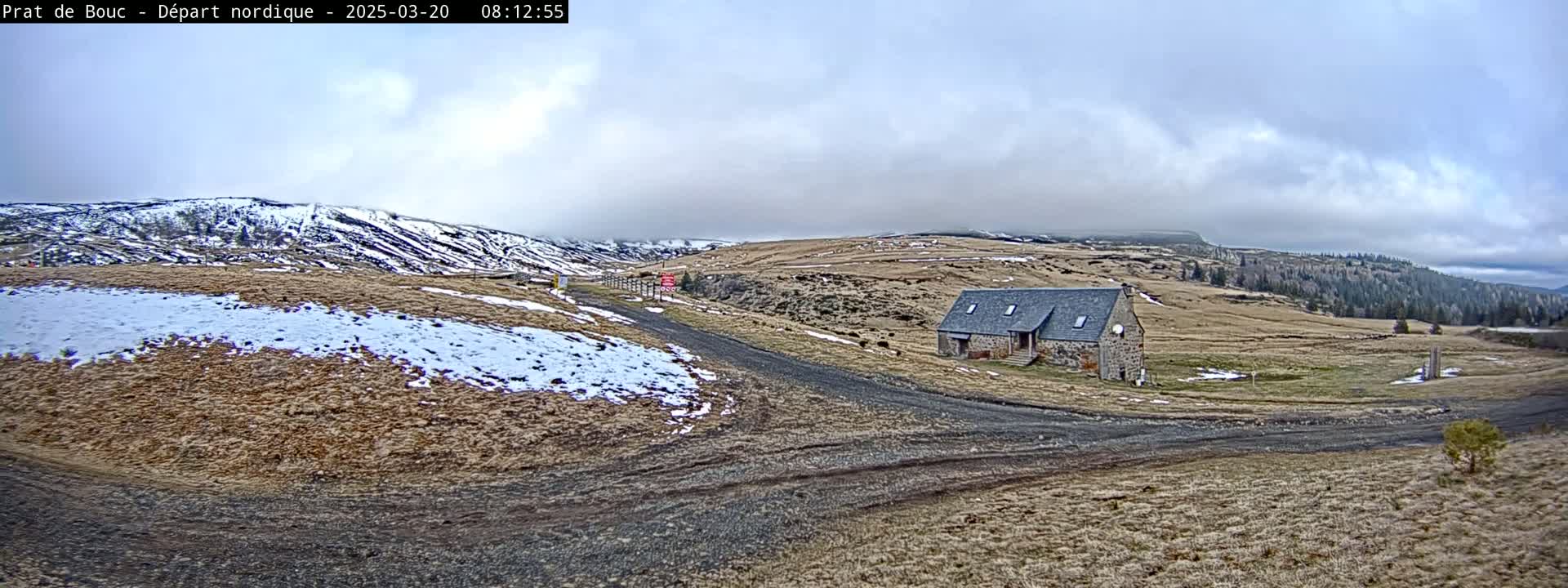 A stone building sits on a gravel road in a hilly, sparsely vegetated landscape with patches of snow under a cloudy sky.