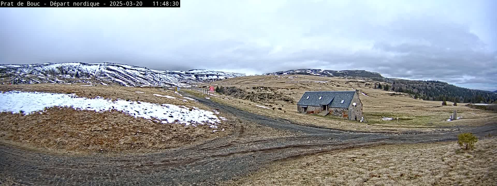 A stone building sits on a dirt road in a sparsely vegetated landscape under a cloudy sky, with patches of snow on the ground and distant snow-covered hills.