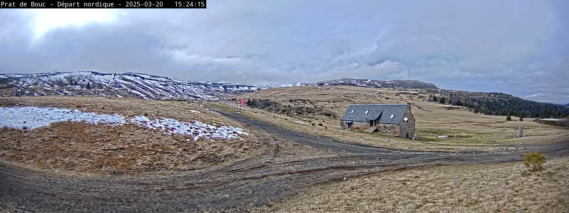 A stone building sits on a dirt road in a sparsely vegetated landscape under a cloudy sky, with snow patches visible on the ground and distant hills.