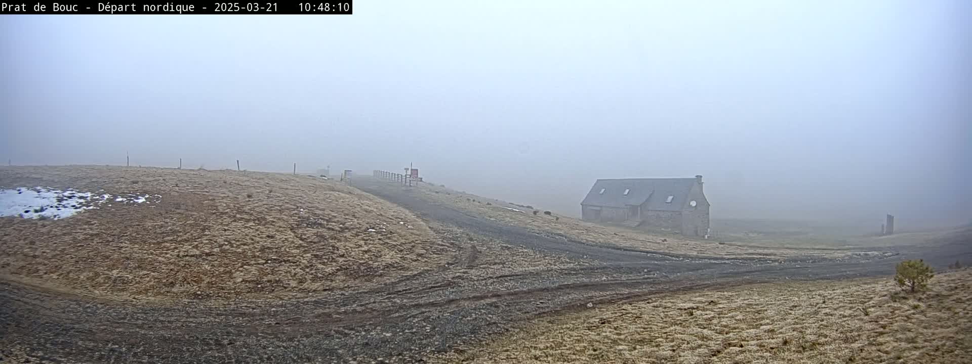 A long, low stone building is visible through a dense fog on a dry, grassy landscape with a dirt road leading to it.