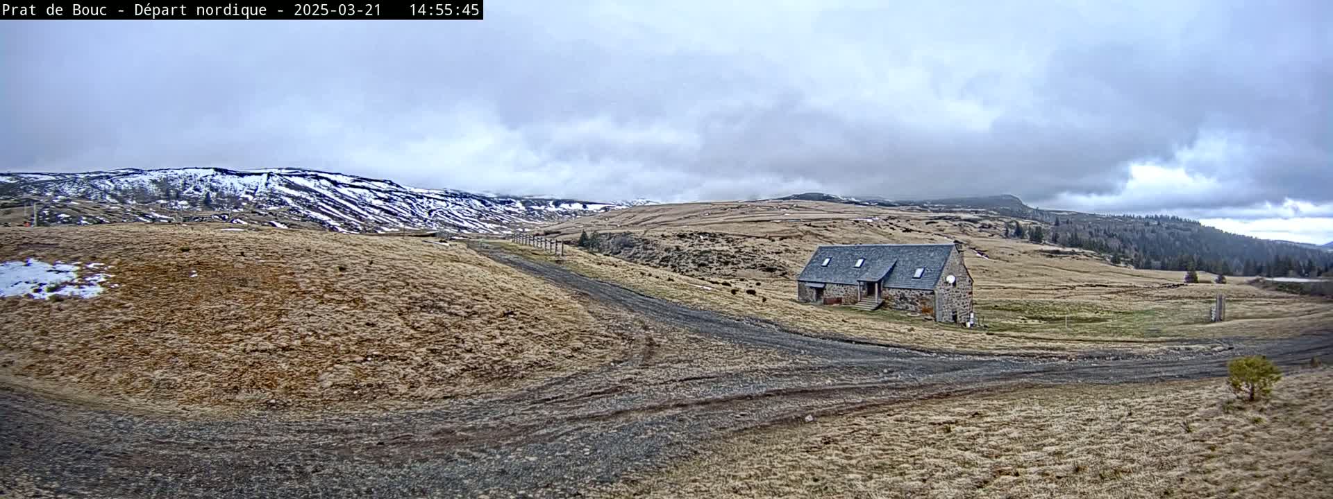 A stone building sits on a dirt road in a mountainous landscape under a cloudy sky, with some snow visible on the distant hills.