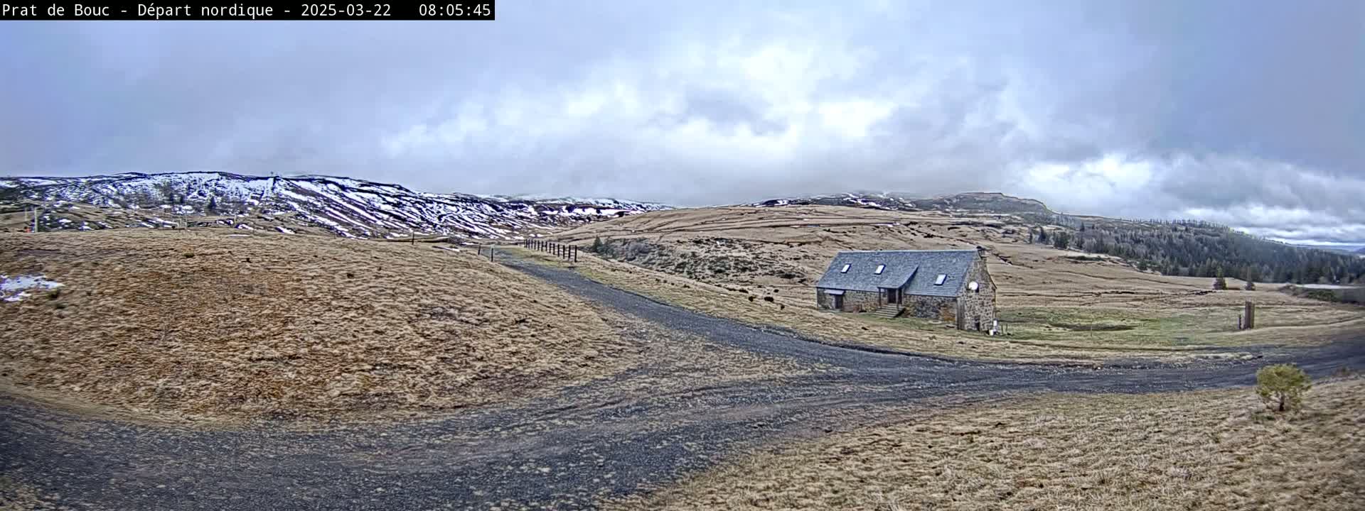 A gravel road leads to a stone building situated in a brown, grassy field under a cloudy sky, with snow-capped mountains in the background.