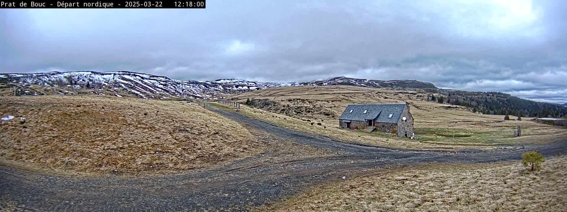 A stone building sits on a dirt road in a dry, hilly landscape under a cloudy sky, with snow-capped mountains in the distance.