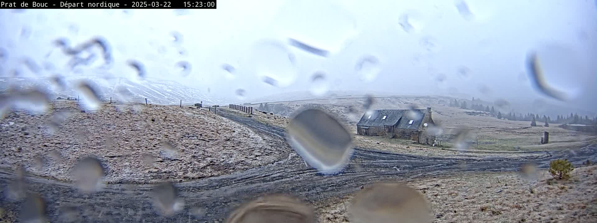 A stone building sits on a snow-dusted hillside viewed through a rain-covered window, with a dirt road leading to it under a cloudy sky.