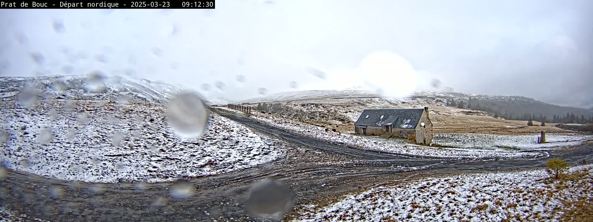 A light snow falls on a dirt road leading to a stone building in a hilly, sparsely vegetated landscape.