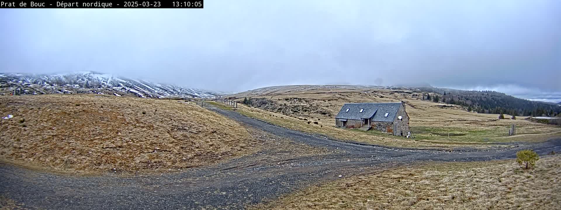 A stone building sits on a gravel road in a hilly, dry landscape under an overcast sky, with snow-covered mountains in the background.