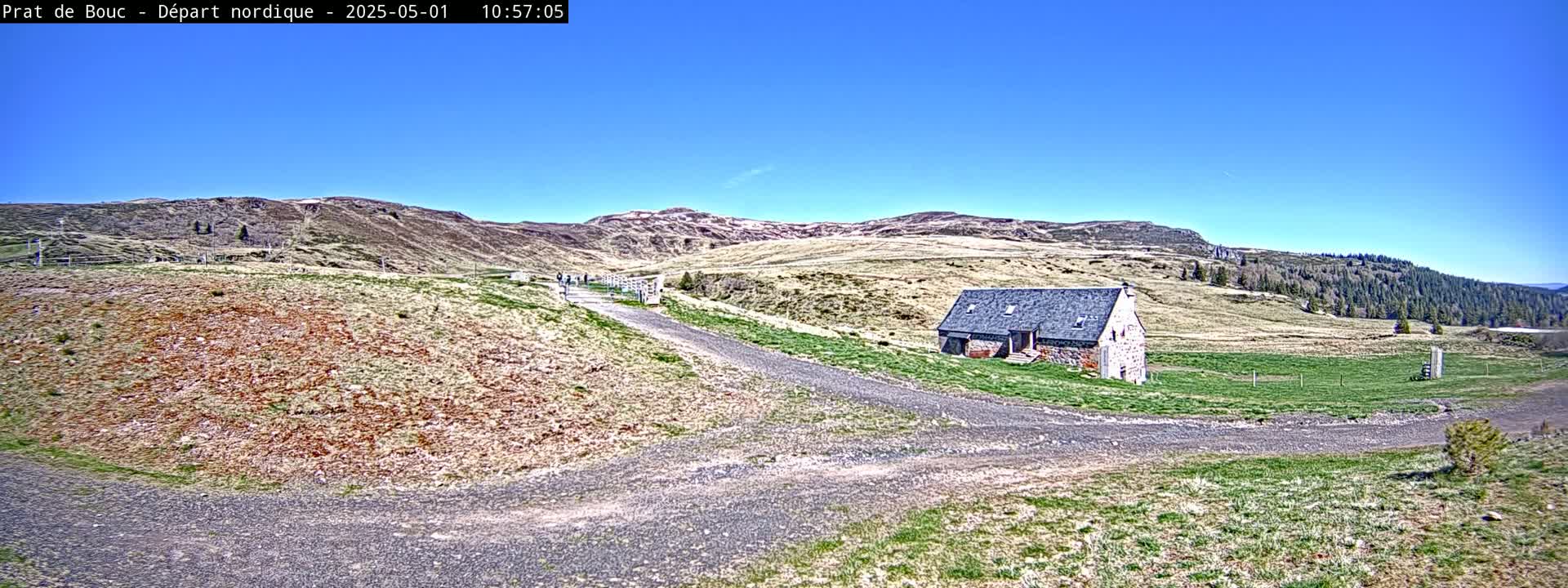 A stone building sits on a grassy hillside next to a gravel path under a clear blue sky.