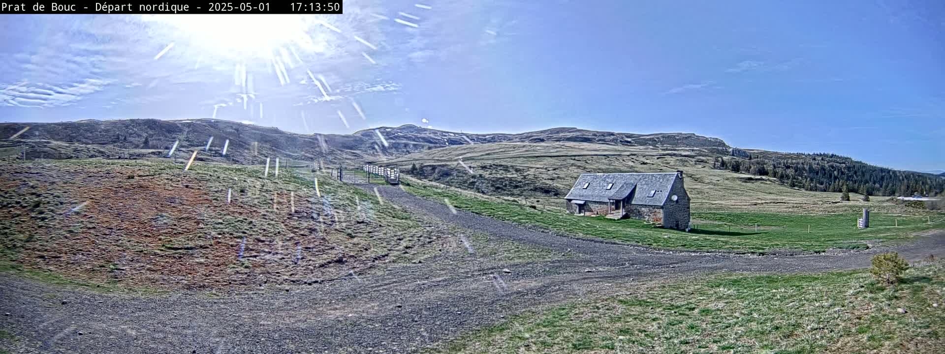 A stone building sits on a grassy hillside under a mostly sunny sky with some light snow falling.