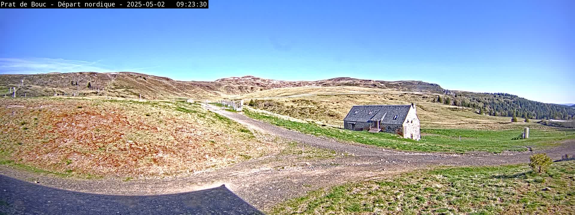 A stone building sits on a grassy hillside with a gravel road leading to it, under a clear blue sky.