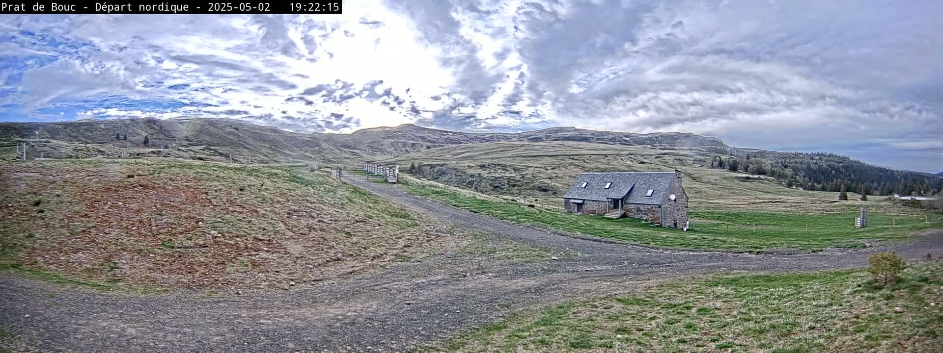 A stone building sits on a grassy plateau under a partly cloudy sky, with a gravel road leading to it and hills in the background.