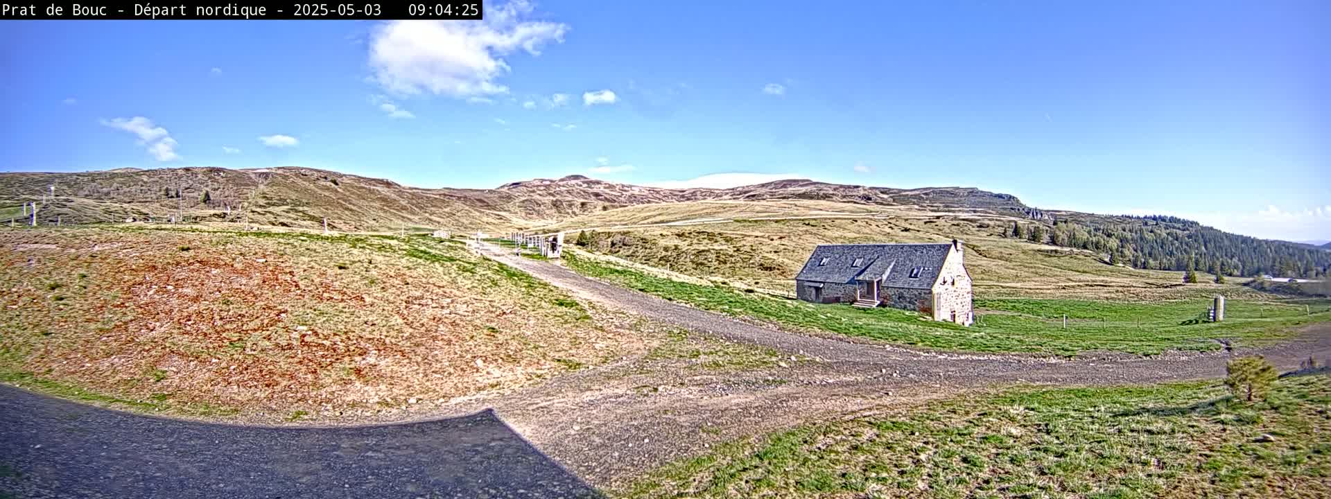 A stone building sits on a grassy hillside near a gravel road under a mostly sunny sky.