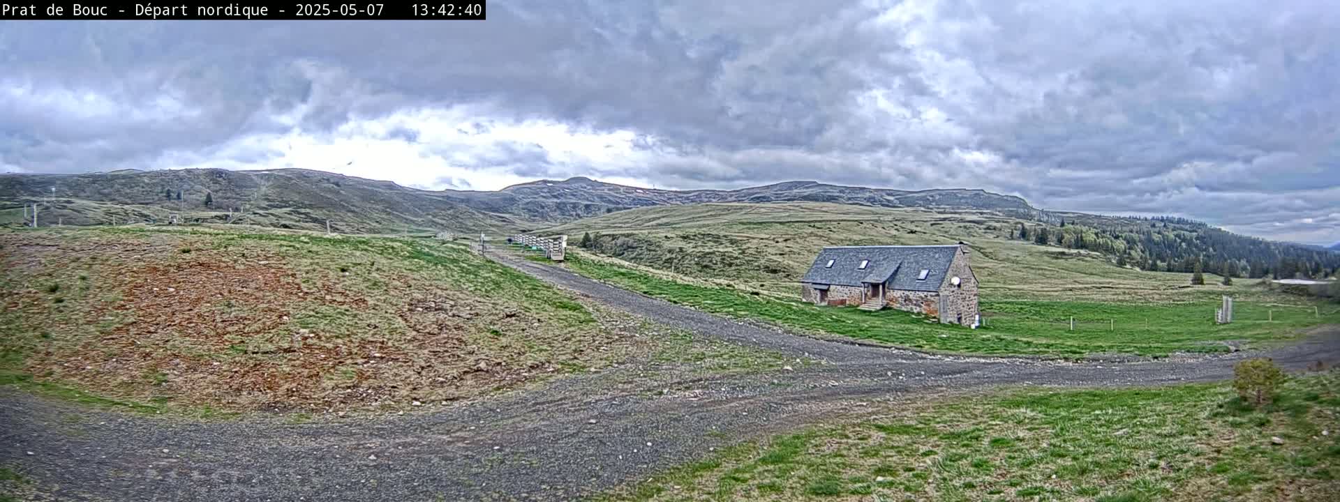 A stone building sits on a grassy hillside, with a gravel road leading to it, under a cloudy sky.