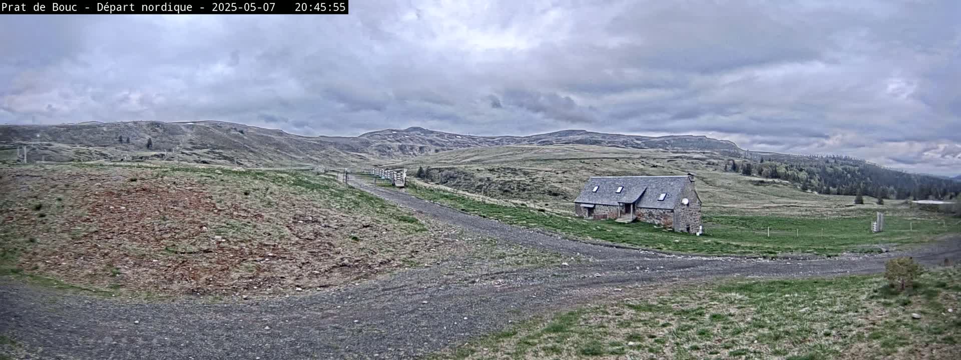 A gravel road leads to a stone building nestled in a hilly landscape under a cloudy sky.