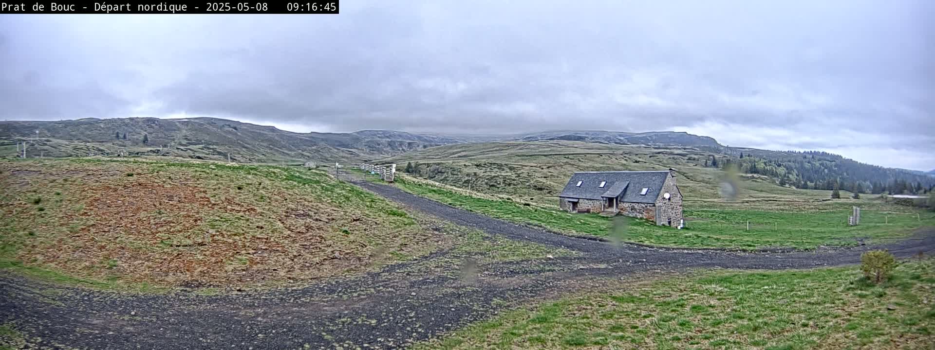 A stone building sits on a grassy hillside under an overcast sky, with a gravel road leading toward it.