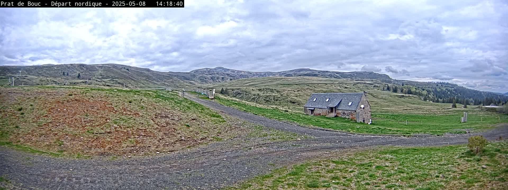 A stone building sits on a grassy plateau, with a gravel road leading toward it, under a cloudy sky.