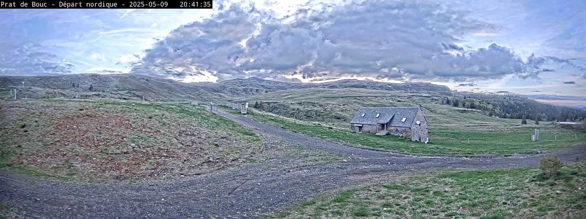 A stone building sits on a grassy hillside, with a gravel road leading to it under a partly cloudy sky.