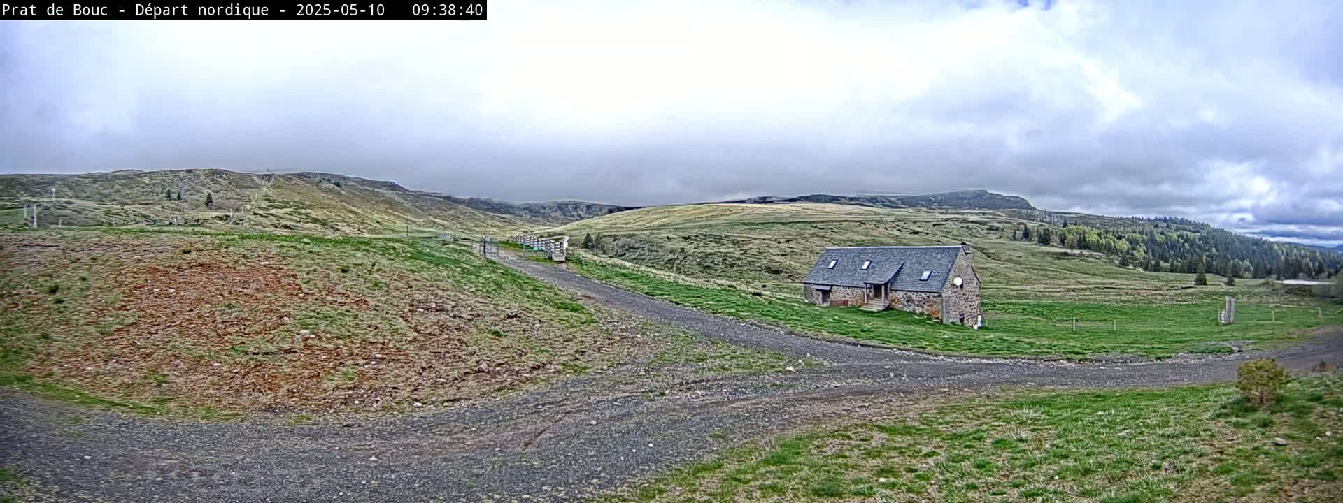 A stone building sits on a grassy hillside, with a gravel road leading to it under a cloudy sky.