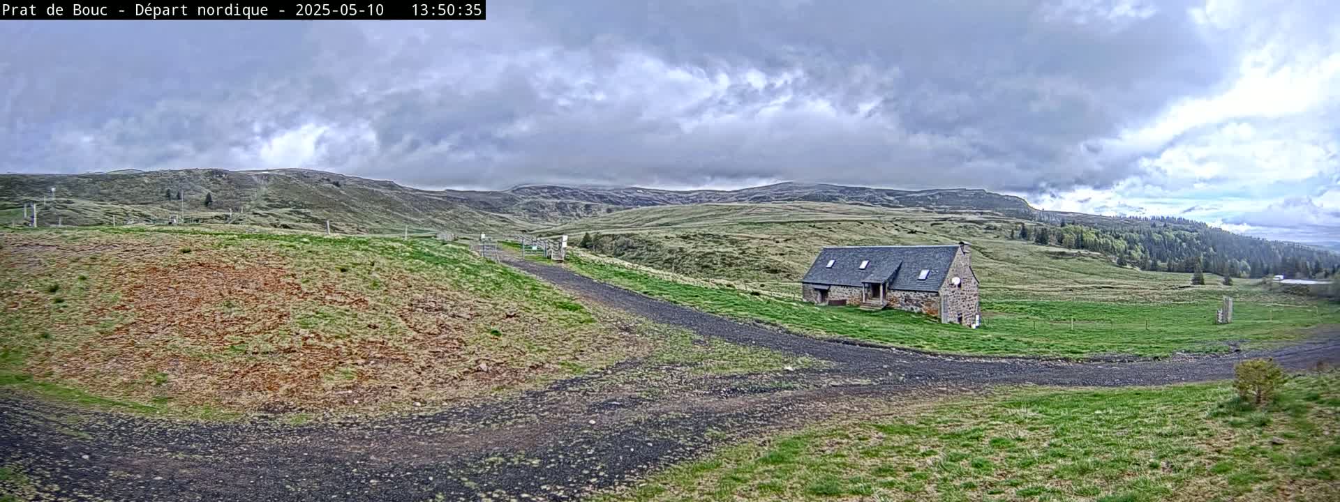 A stone building sits on a grassy hillside under a cloudy sky, with a gravel road leading to it.