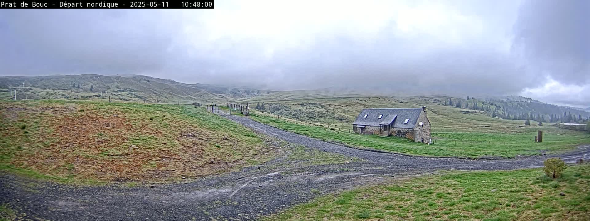 A stone building sits on a grassy hillside, with a gravel path leading toward it under a cloudy sky.