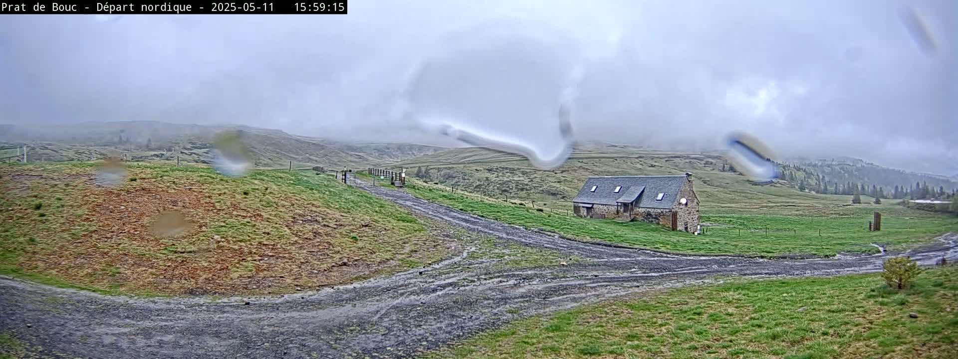 A stone house sits on a grassy hillside in the rain, with a muddy track leading toward it from a higher vantage point.