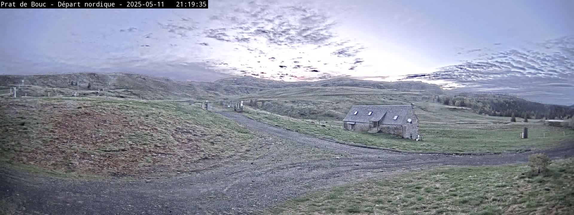 A gravel road leads to a stone building in a hilly, grassy landscape under a cloudy, twilight sky.
